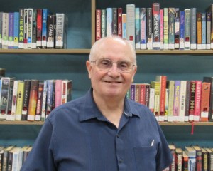 A man wearing glasses and a blue shirt stands in front of books on a shelf